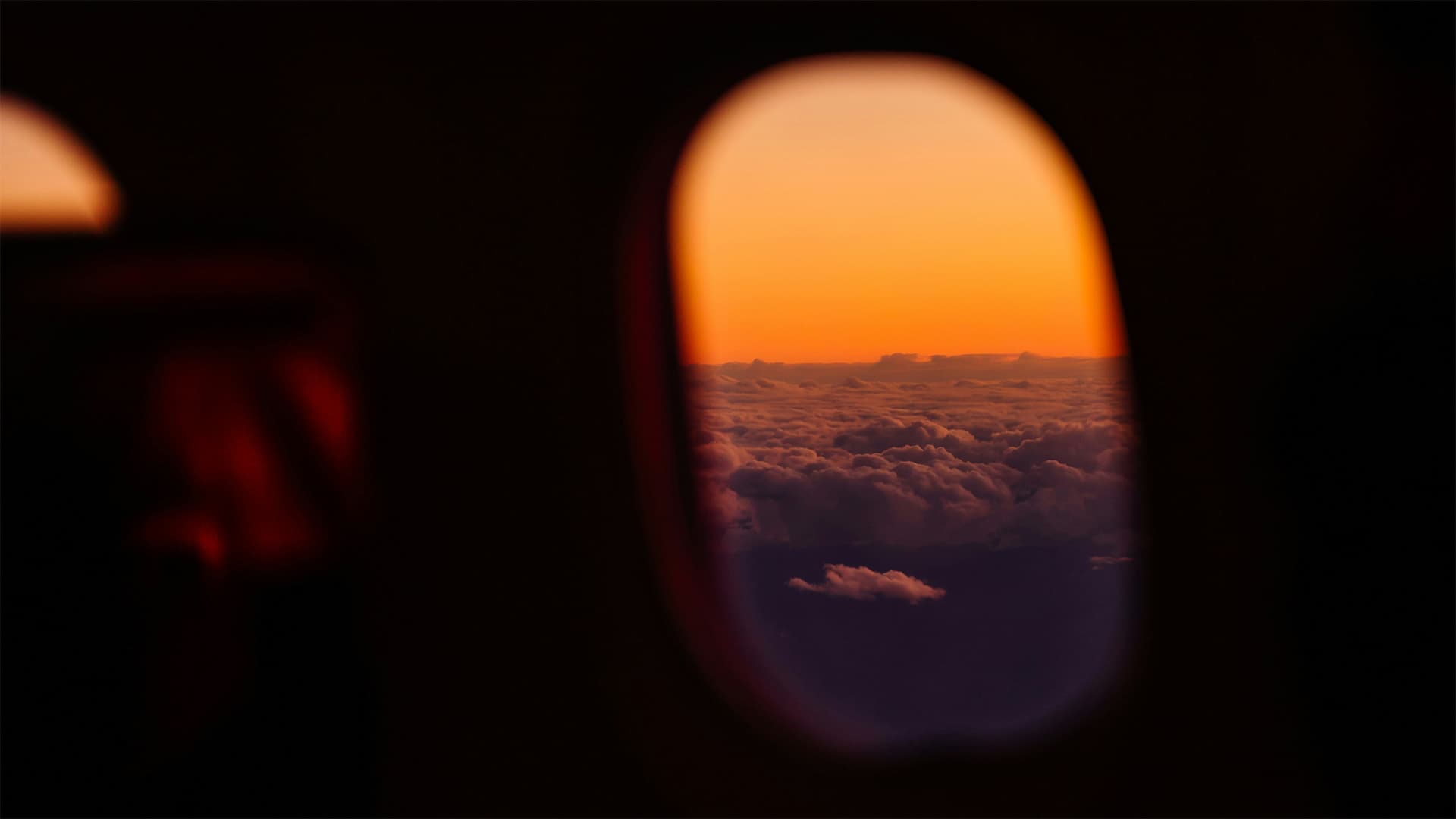 Utsikt genom ett flygplansfönster över ett hav av moln vid solnedgång, med en orange himmel i horisonten.