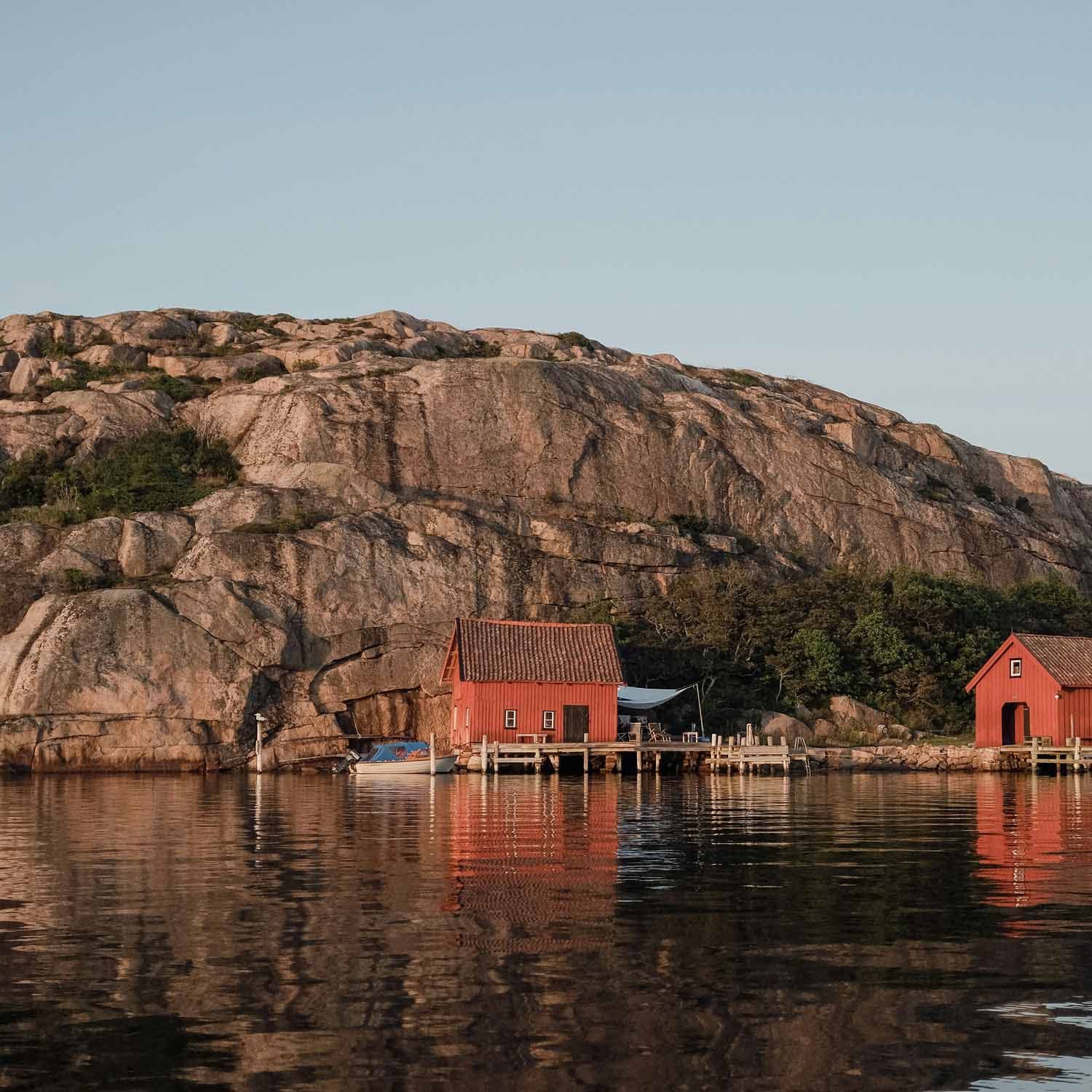 Röda sjöbodar vid vattnet framför klippor i en kustmiljö på västkusten.